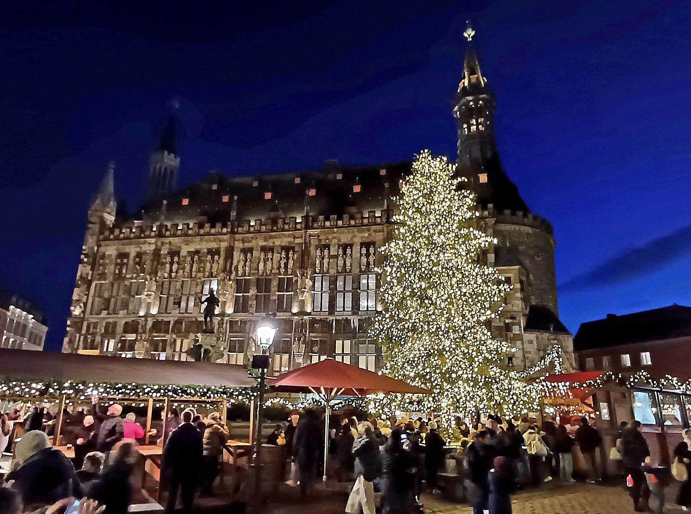 A lit Christmas tree stands before Aachen town hall as the Christmas market fills the square at night