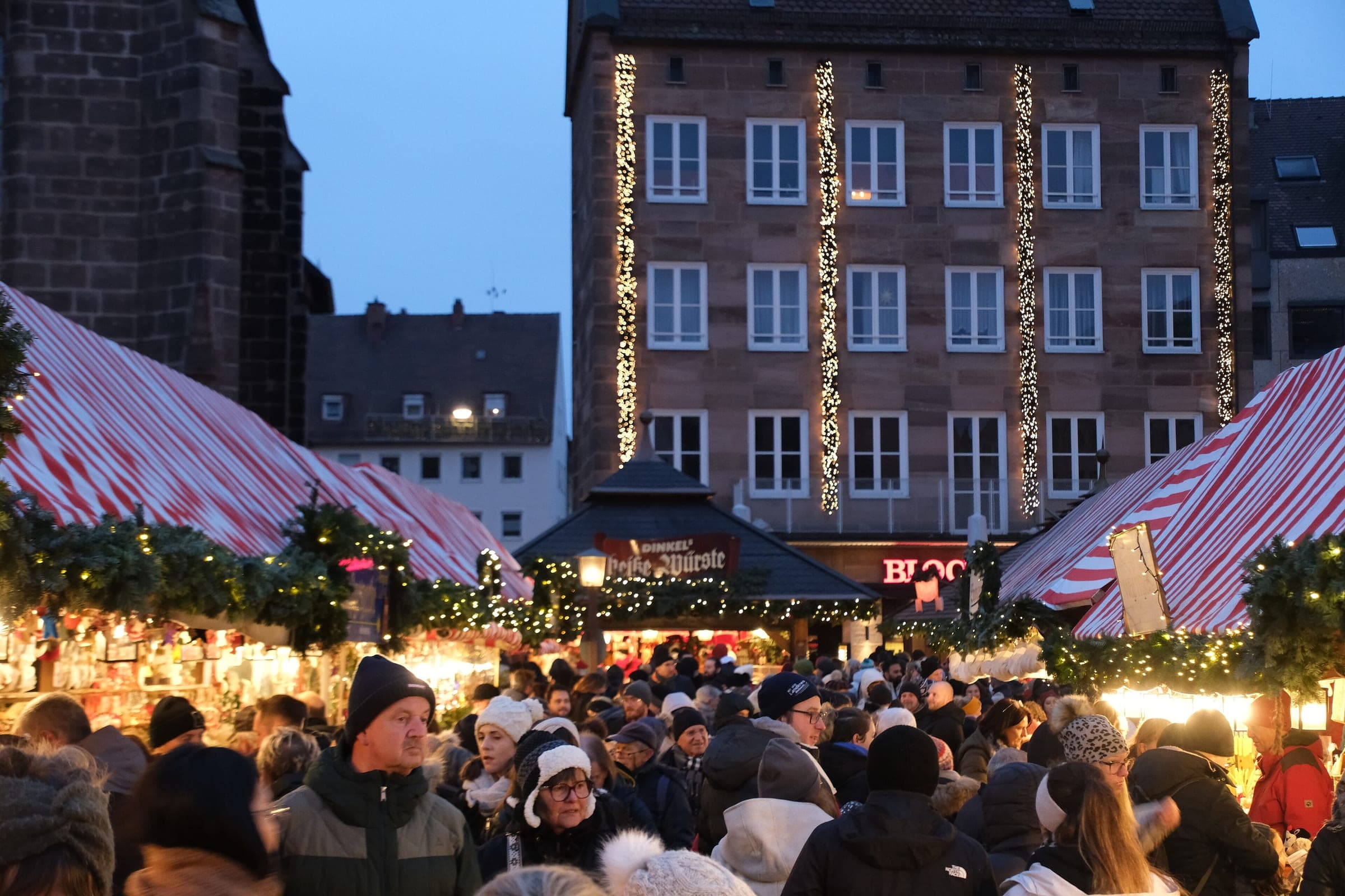 A crowd fills a lane of garlanded stalls at the Nuremberg Christkindlesmarkt below a building strung with lights