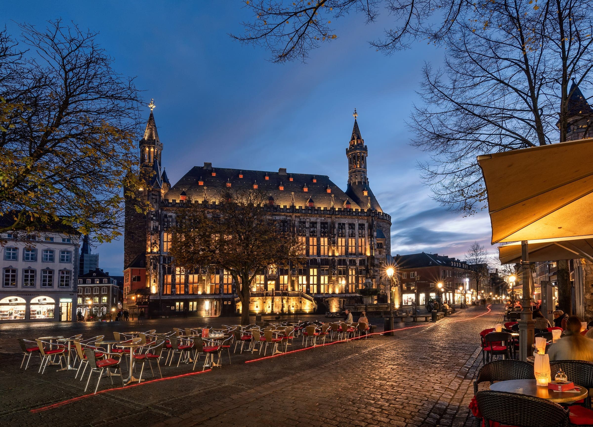 Aachen town hall glows at blue hour across the Markt with café tables set out on the cobbles