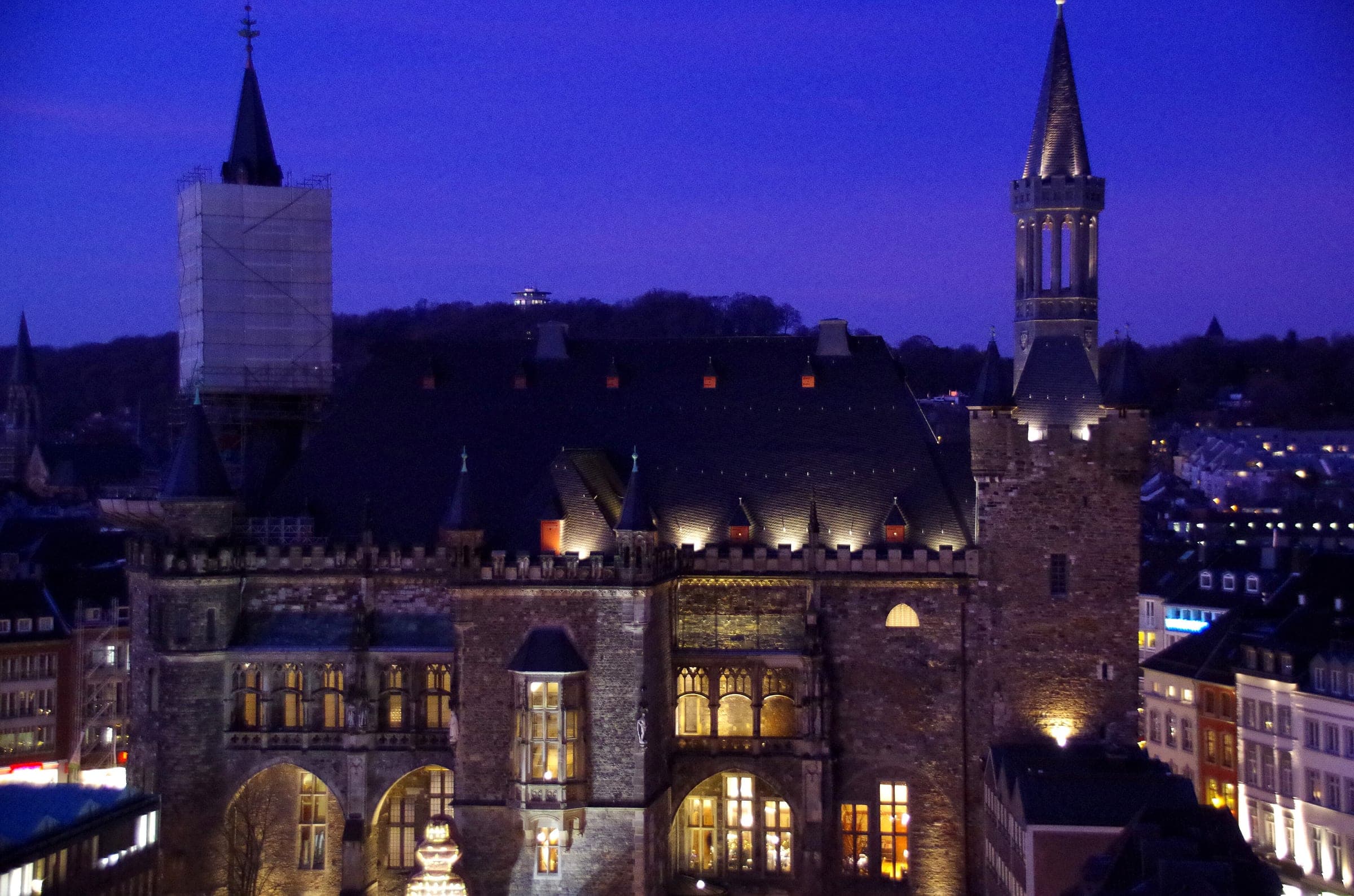 Lit windows and towers of Aachen town hall stand against a violet evening sky
